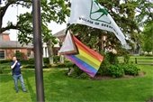 Photo of a man raising the Pride flag below the Village flag in front of Village Hall