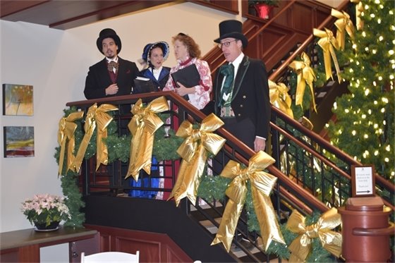 Photo of a choir in Dickens style dress performing while standing on the stairs at Village Hall in front of a crowd